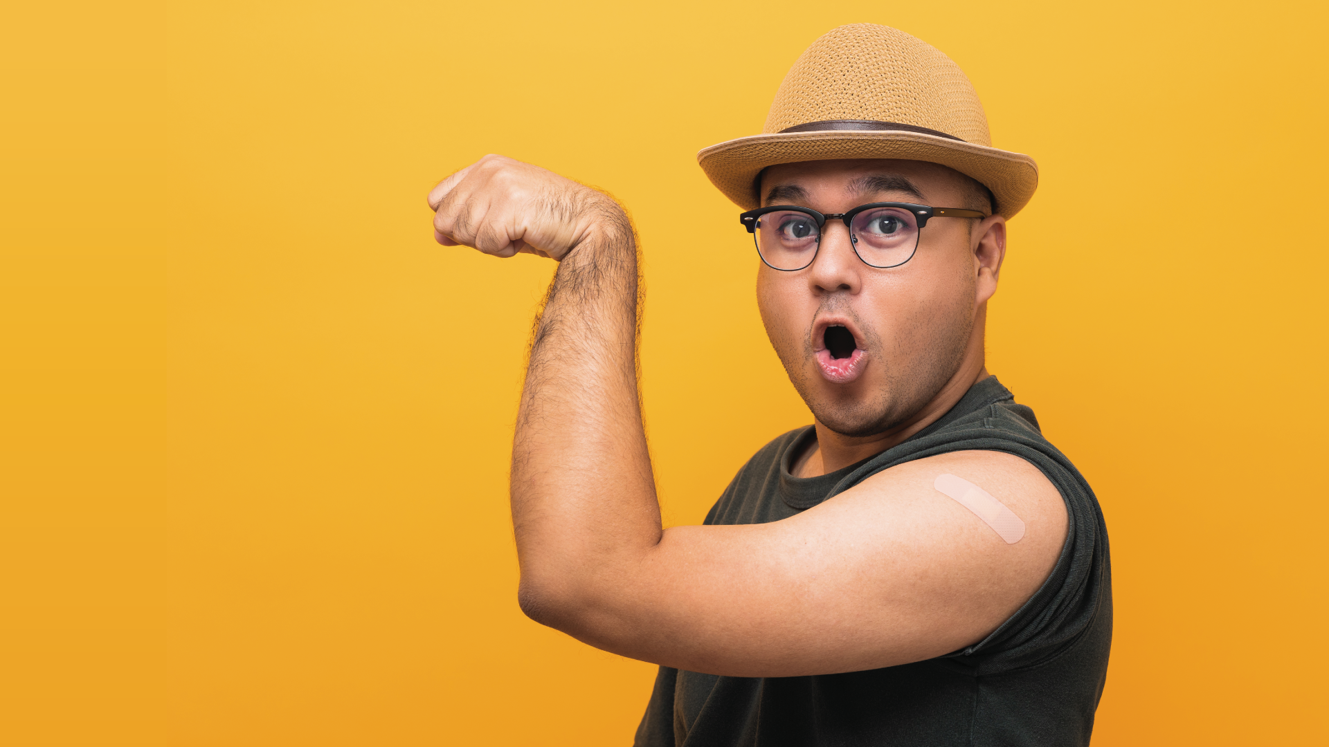 young man with glasses and straw hat makes a silly expression while showing his muscle and a bandaid from a vaccine