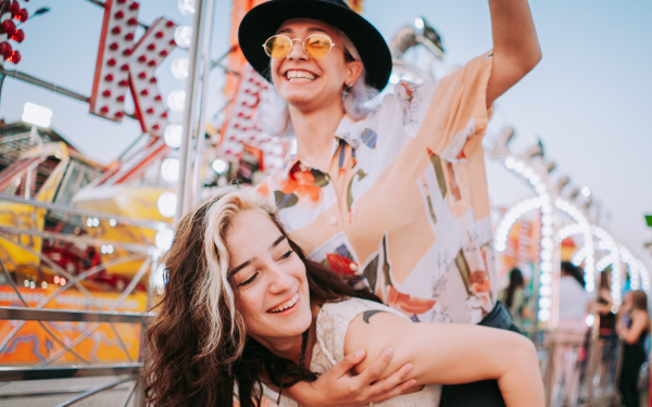 Two teenagers laughing as one carries the other on their back at a carnival.