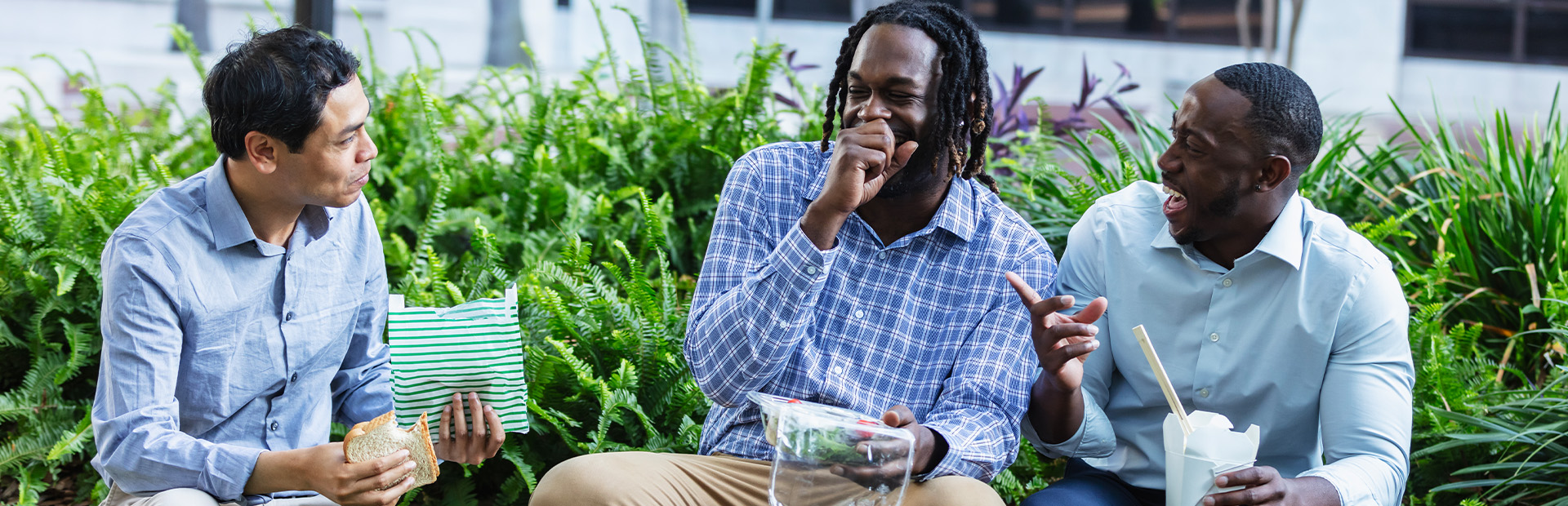 Three men sitting outdoors, eating lunch and laughing together.
