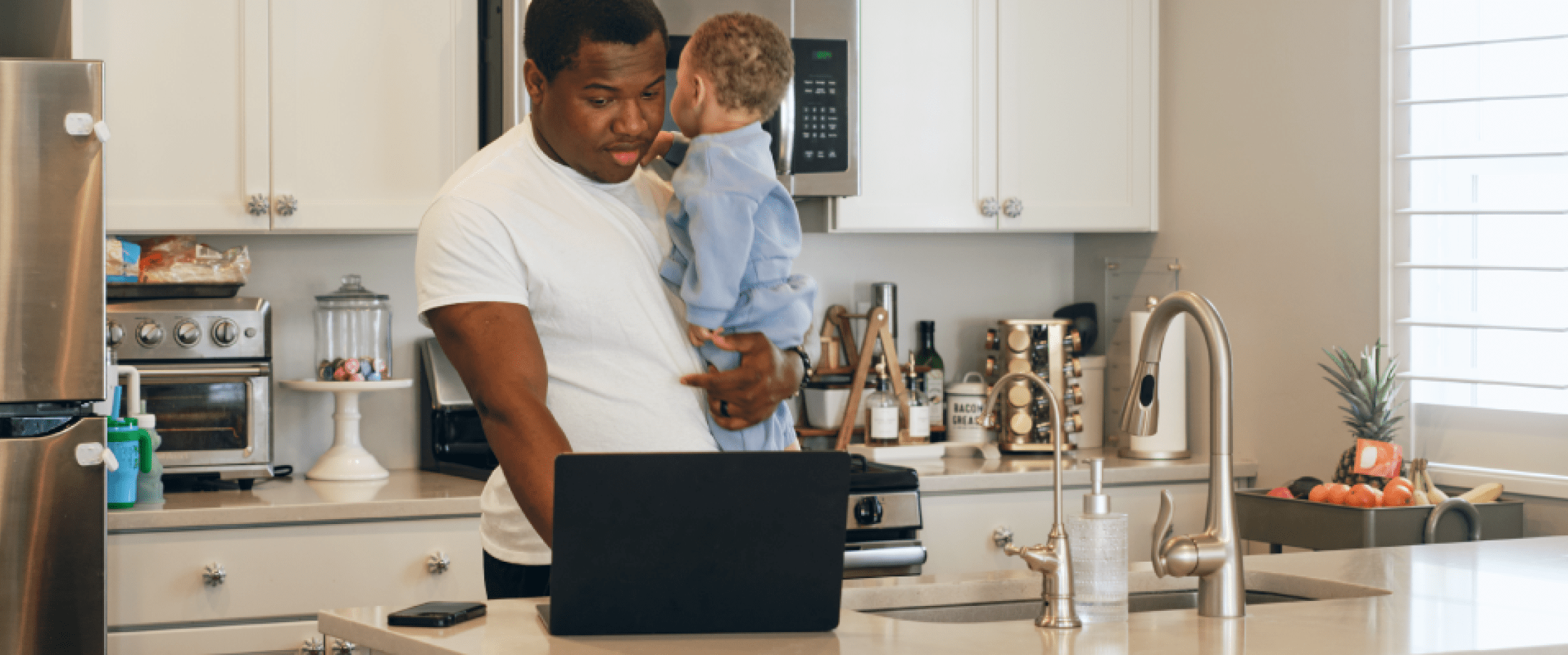 Father holding infant while working on a laptop in the kitchen