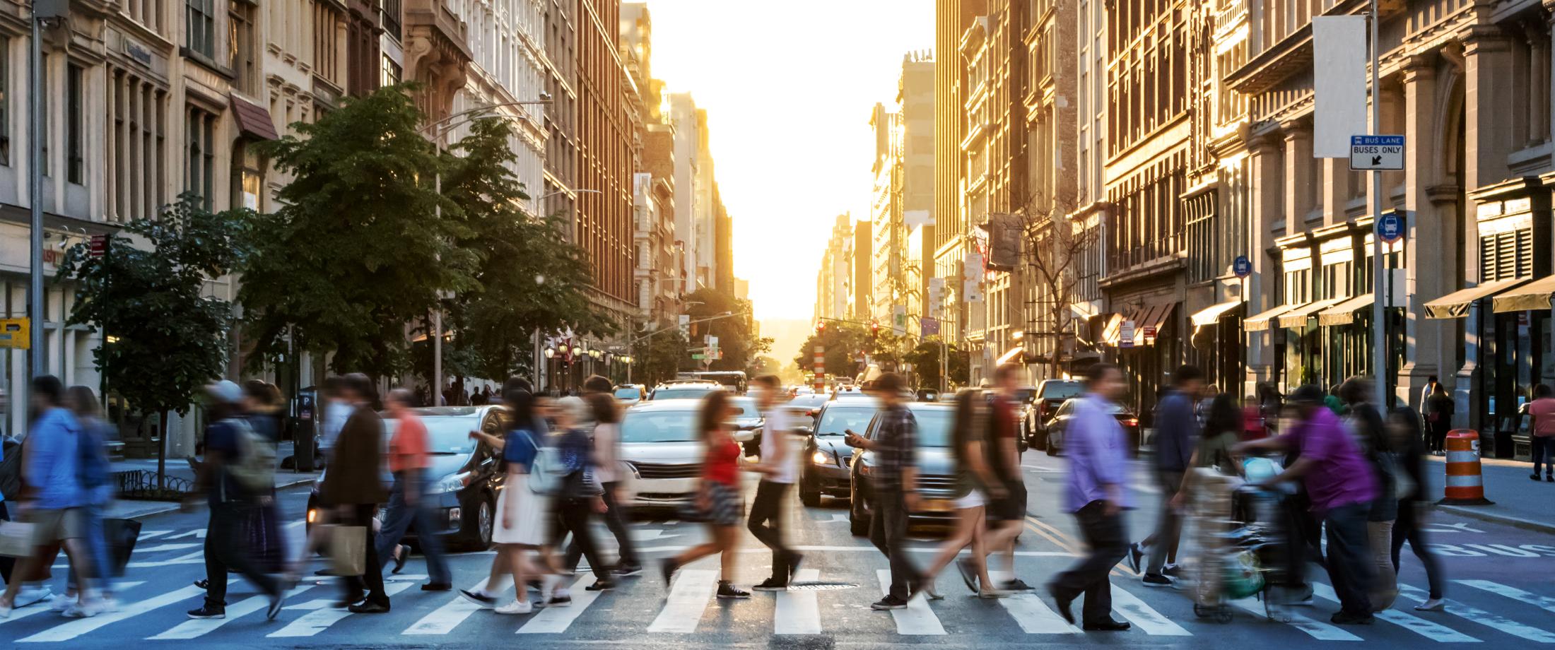Many people crossing a city street at sunset.