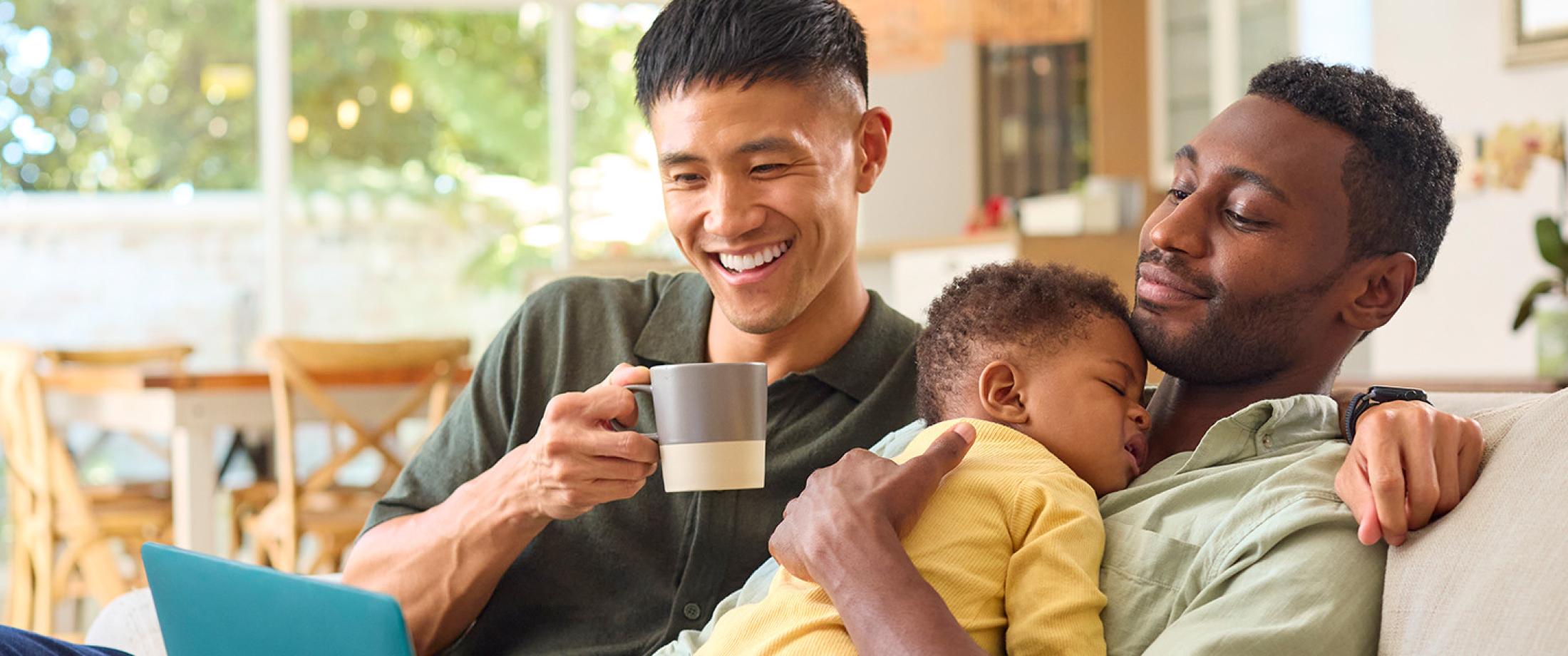 Two parents sitting on a couch while one holds a sleeping baby.