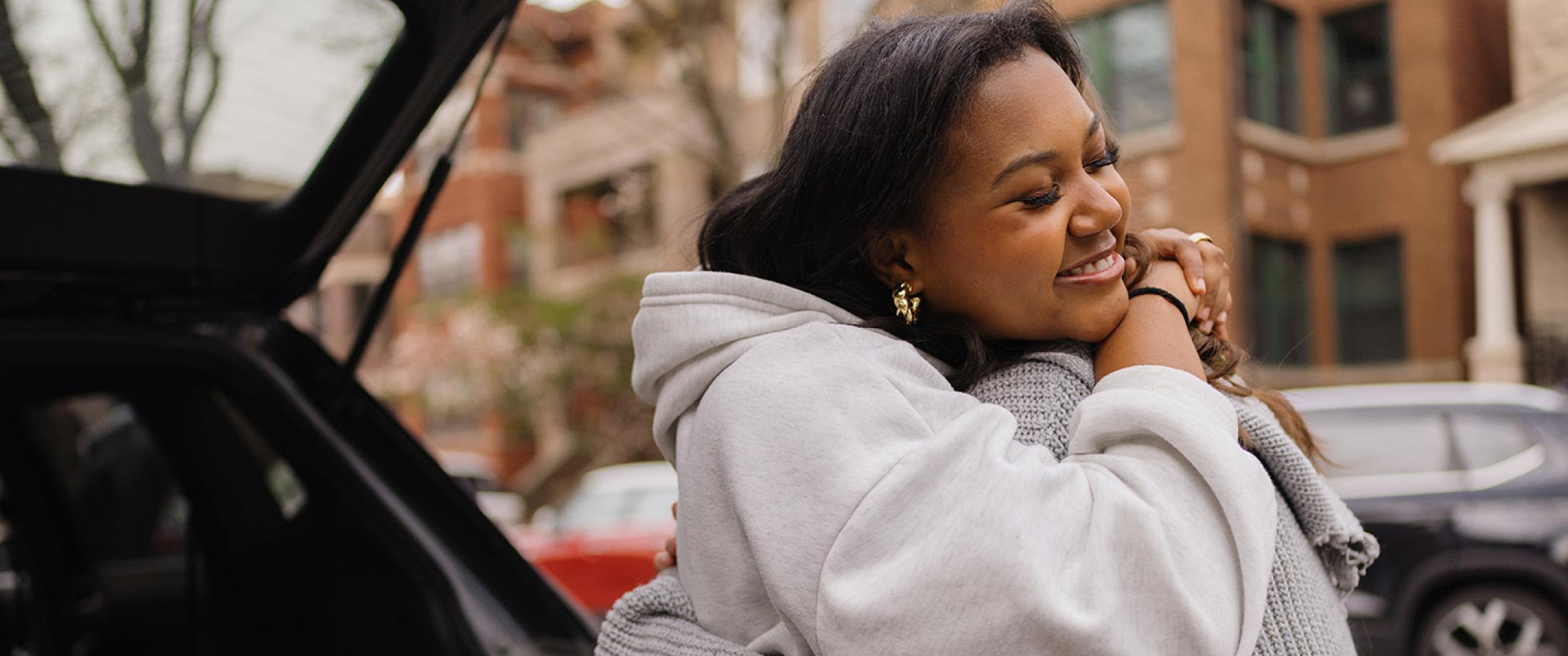 Two women hugging near a parked car on a residential street.