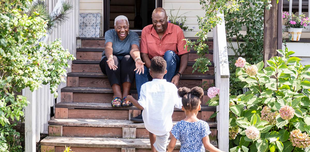 Two older adults sitting on a porch smiling as two children walk toward them.