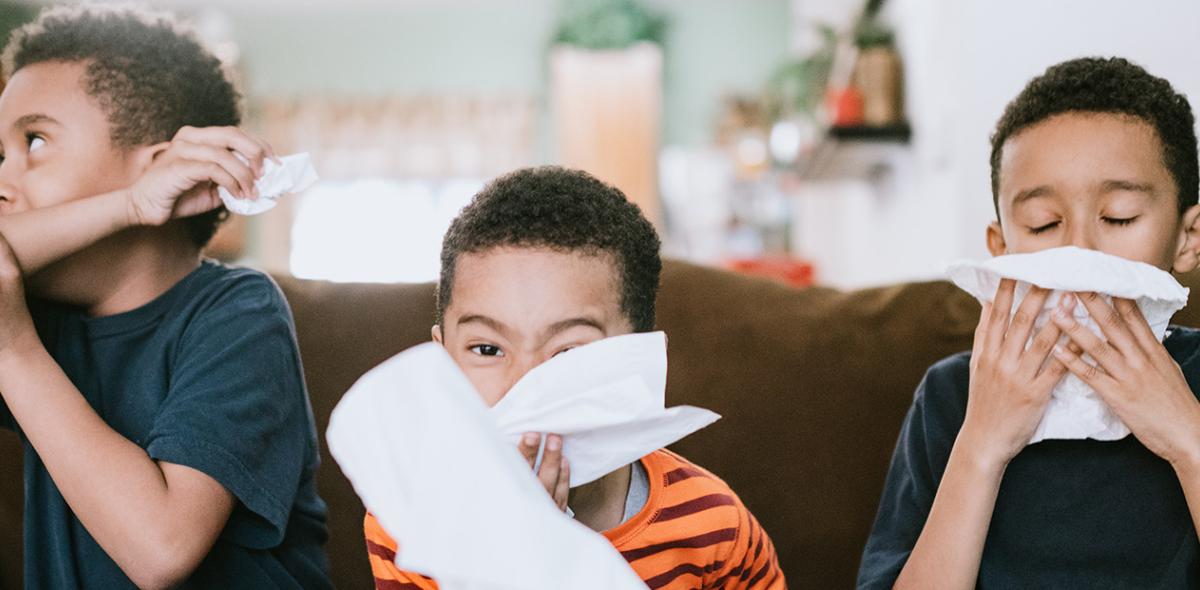 Three children sitting on a couch using tissues to wipe their noses.