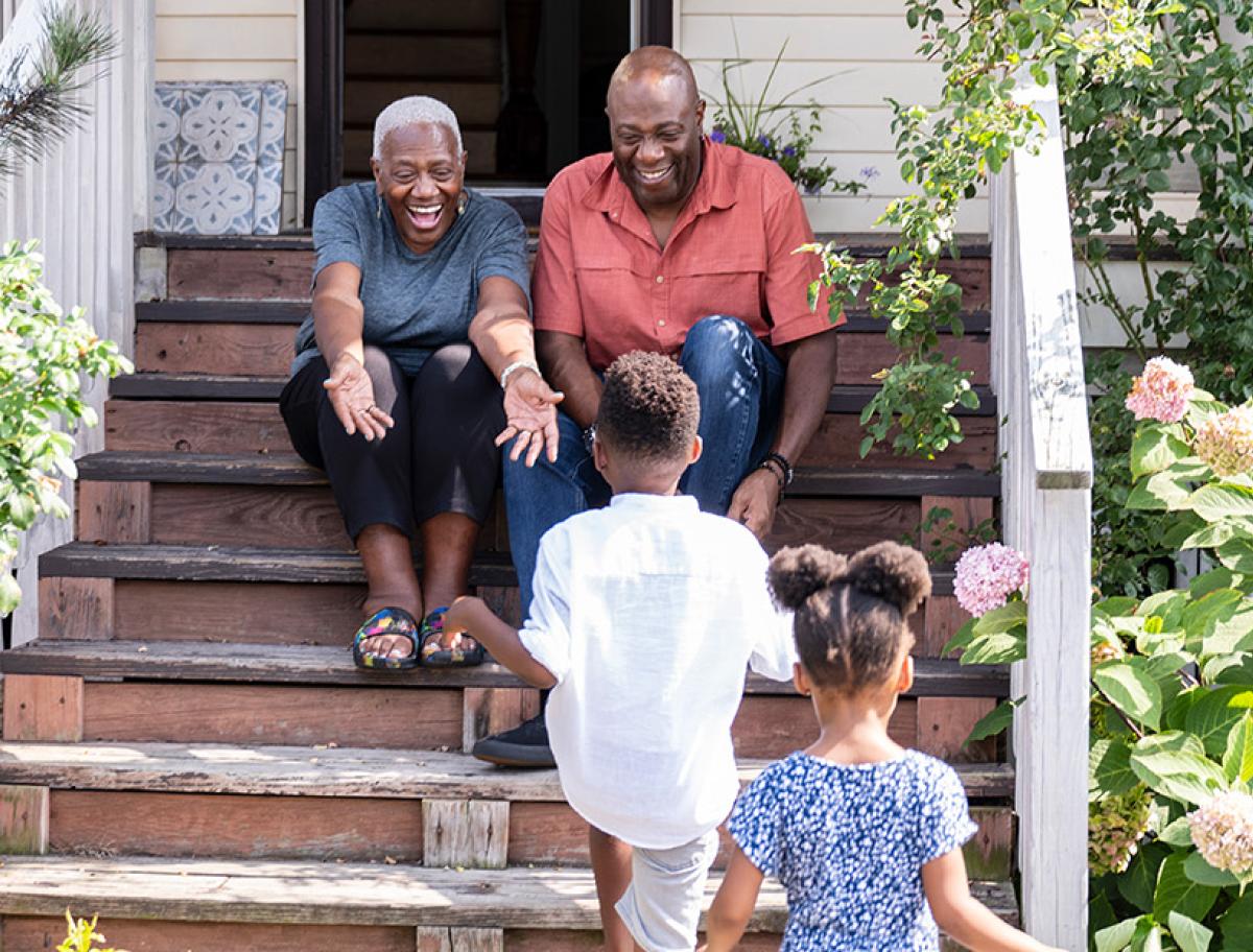 Two older adults sitting on a porch smiling as two children walk toward them.