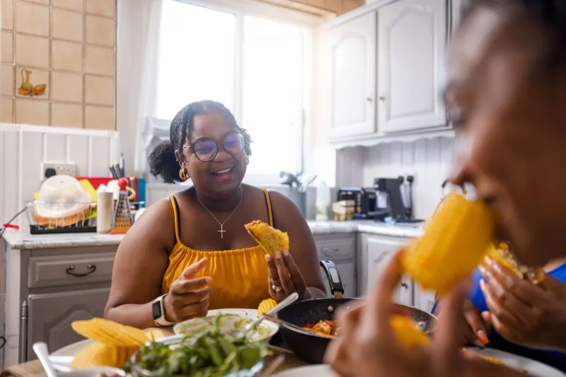 Laughing woman eating corn on the cob with a friend at the kitchen table