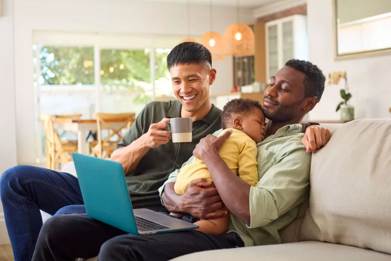 Male couple sitting on couch holding their baby and looking at a laptop together
